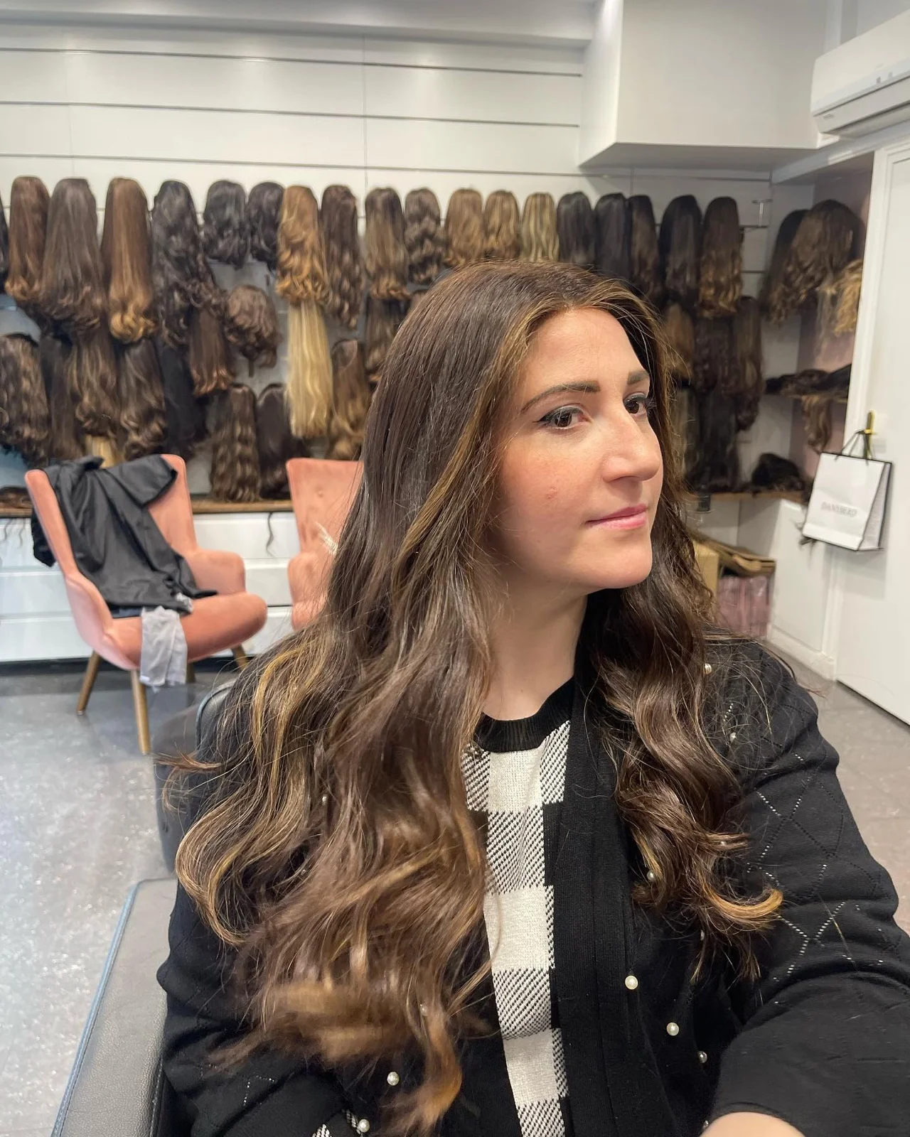 Cliente de profil devant le mur de perruques en cheveux naturels du salon, au moment du choix