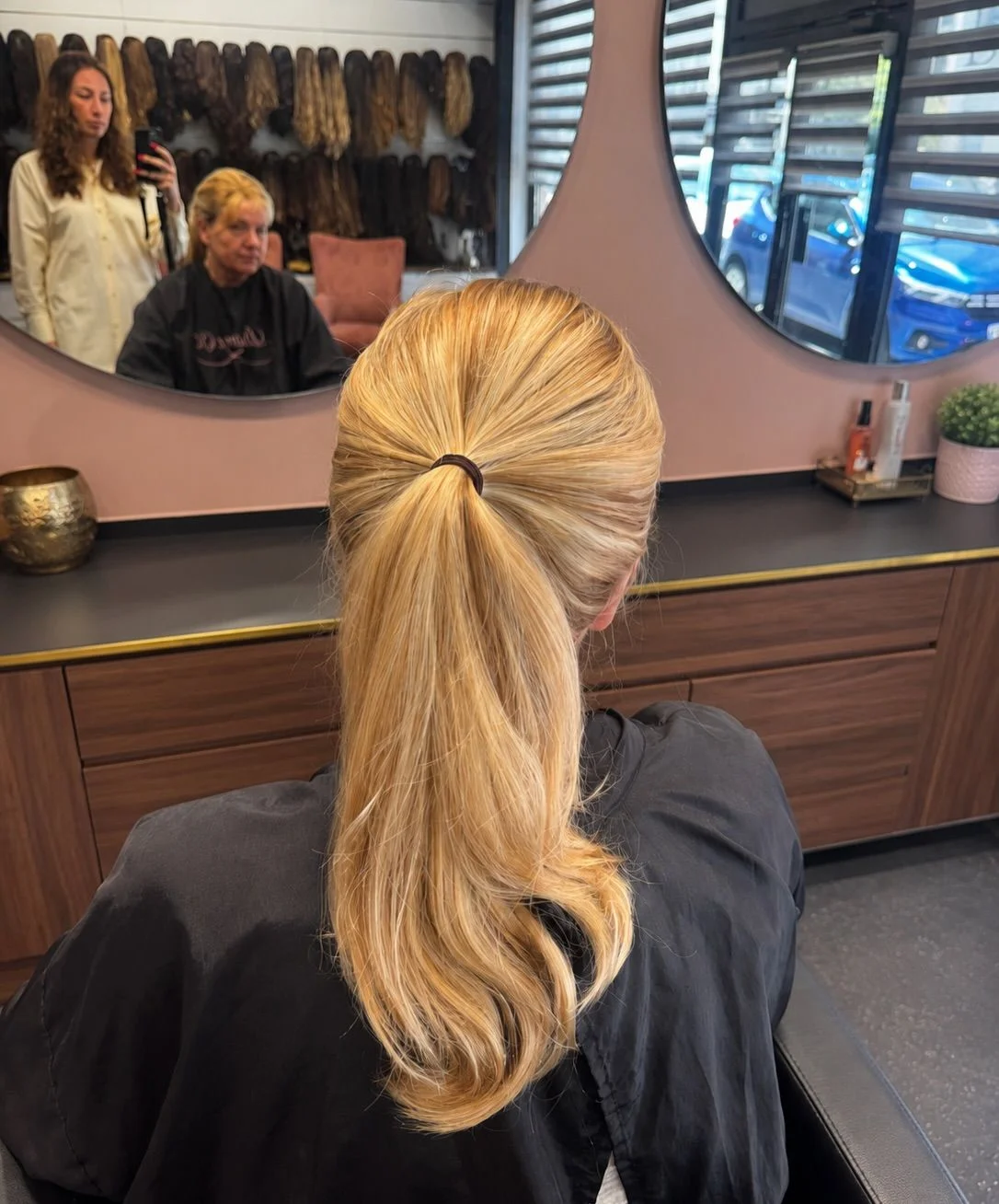 Perruque blonde coiffée en queue de cheval basse, vue de dos devant le miroir du salon, Dvora K