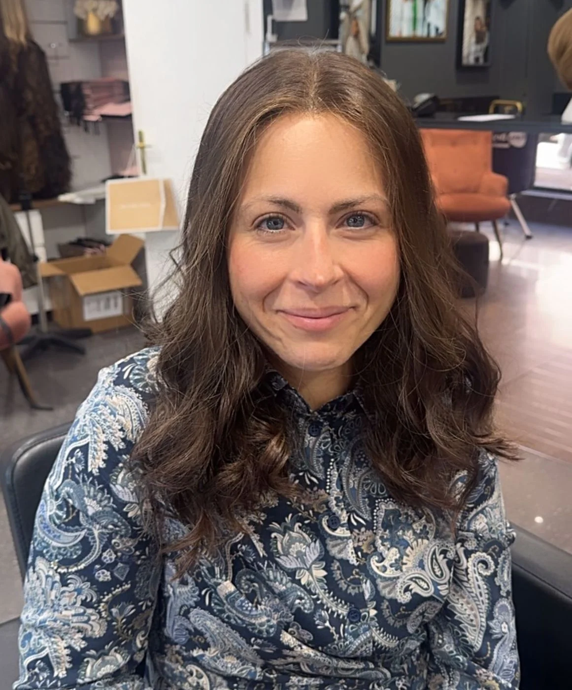 Cliente souriante portant une perruque mi-longue brune légèrement ondulée, assise au salon